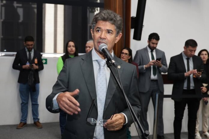A man in a gray suit speaks into a microphone at a press event, while colleagues stand behind him.