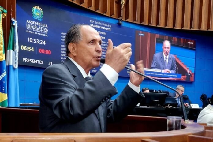 Man in a suit speaking at a podium in a legislative chamber with a blue backdrop and flag nearby, microphone in front of him.