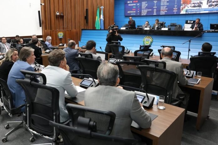 Formal government hearing with officials at desks, camera operator filming, and a blue electronic board behind.