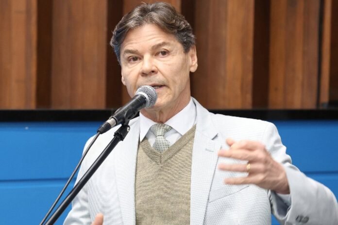Man in a light gray blazer speaks into a microphone during a public event.