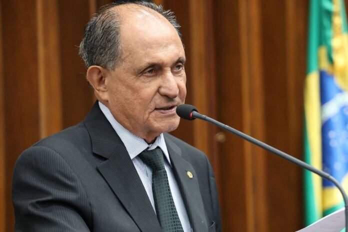 Older man in a dark pinstripe suit speaking into a microphone at a podium, with a Brazilian flag in the background.