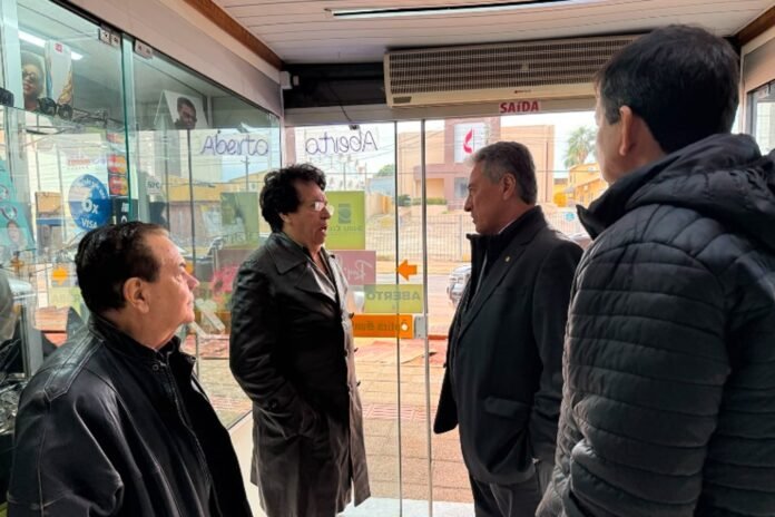 Four adults in jackets stand inside a glass storefront, talking with the woman in the center addressing two men nearby. Outside street visible through glass doors.