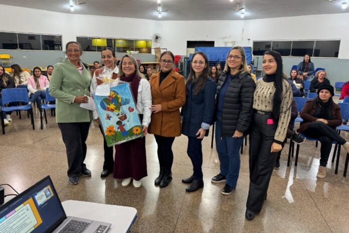 Seven women stand in a meeting hall presenting a colorful handmade quilt featuring flowers and figures while an audience watches nearby.