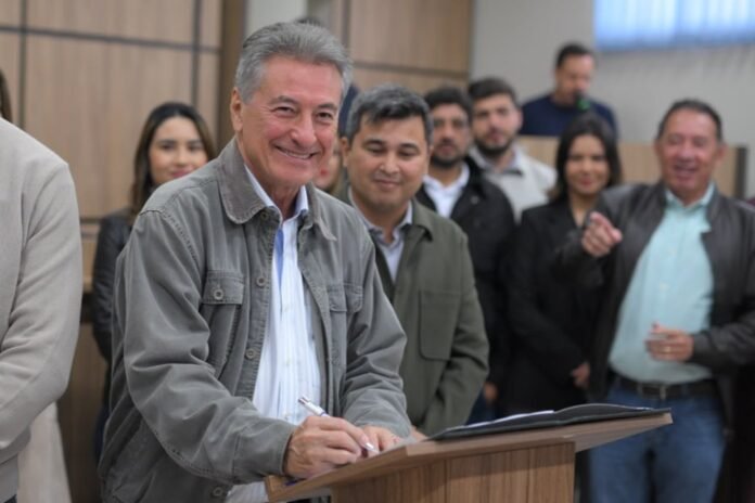 Man in a gray jacket signing a document at a wooden podium as a group of colleagues watch and smile indoors.