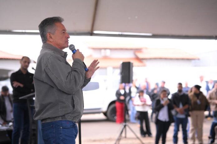 Man in a gray jacket speaks into a microphone at an outdoor event under a tent, with a blurred crowd behind him.