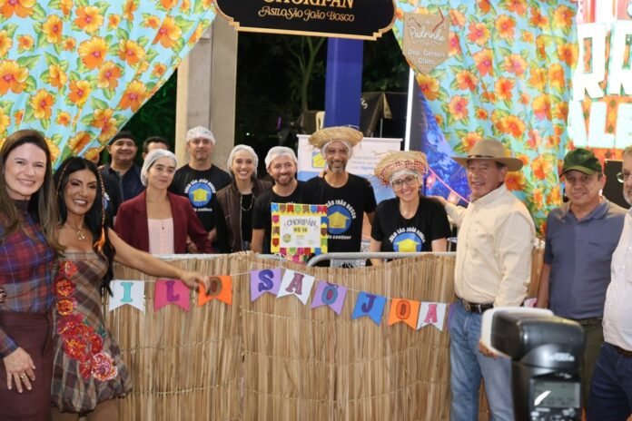 Group of people posing behind a straw fence at a colorful tropical festival booth with floral banners and signs.