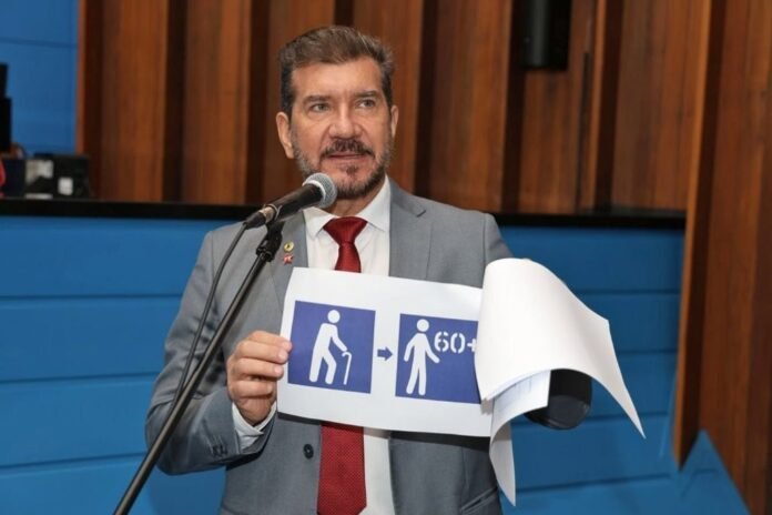 Man in a gray suit and red tie speaks at a microphone while showing a sign with accessibility icons (wheelchair and a person with a cane).