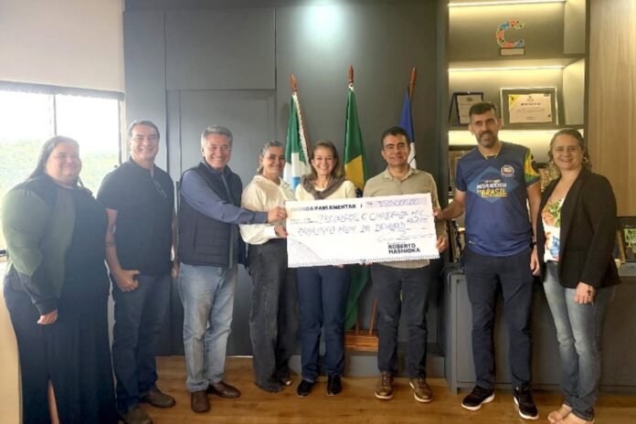 Eight people stand in an office, holding a large ceremonial check in front of flags and a bookshelf backdrop.