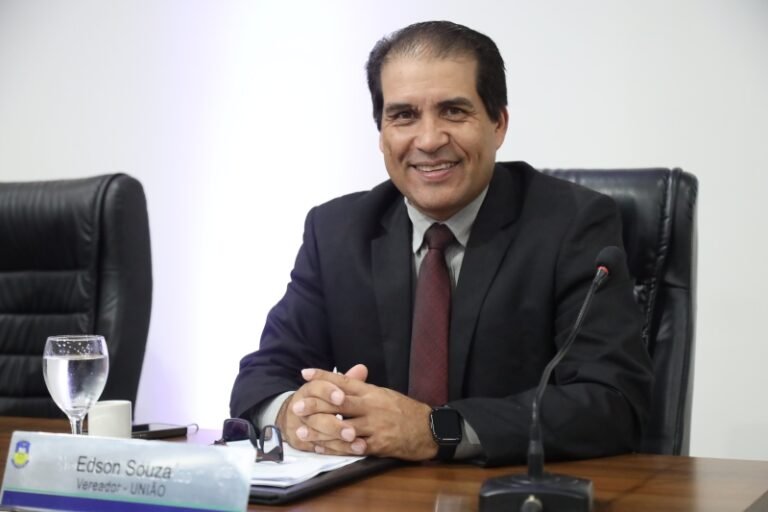Smiling man in a dark suit and red tie sits at a conference table with a microphone in front of him.