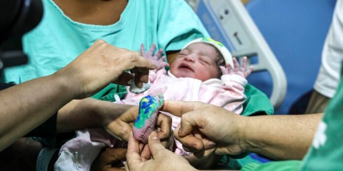 Newborn baby in a hospital bed, surrounded by medical staff as their feet are inked for footprints as a nurse or doctor touches them.