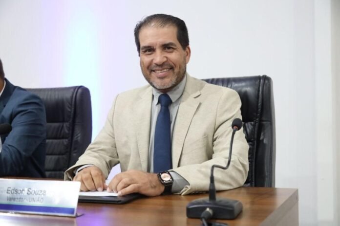 Smiling man in a beige blazer and blue tie sits at a conference table with a microphone and papers ahead.
