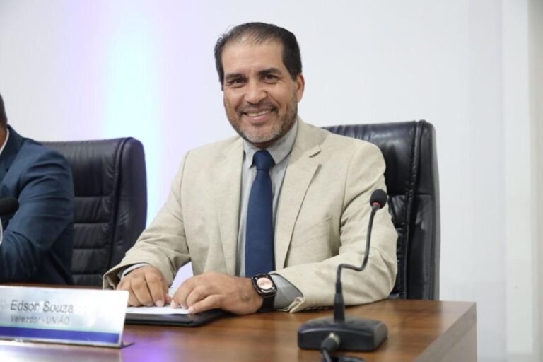 Smiling man in a beige blazer and blue tie sits at a conference table with a microphone and papers ahead.