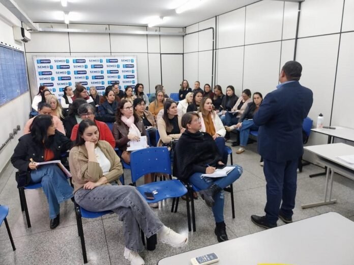 Presenter in a navy suit speaks to a classroom of adults seated in blue chairs at a seminar room with SEMED backdrop behind them