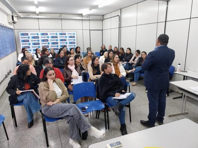 Presenter in a navy suit speaks to a classroom of adults seated in blue chairs at a seminar room with SEMED backdrop behind them