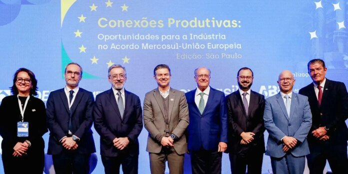 Eight professionals in business attire stand in a row on a stage, posing for a photo in front of a blue backdrop that reads 'Conexões Produtivas: Oportunidades para a Indústria no Acordo Mercosul-União Europeia, Edição: São Paulo' (a conference on trade opportunities).