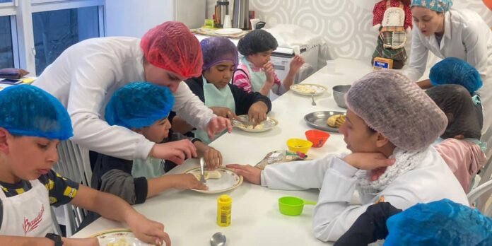 Children wearing hairnets and aprons sit around a long table, guided by adults in white coats as they cut and shape dough for cookies in a bright kitchen classroom.