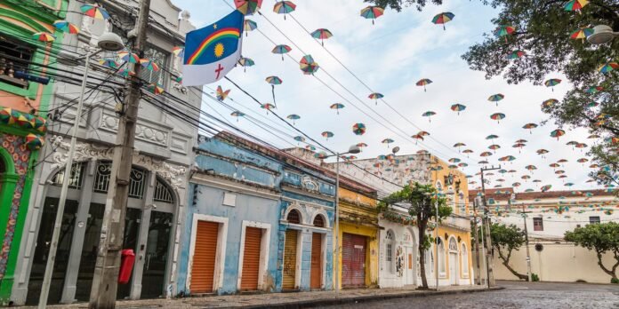 Colorful umbrella decorations span a sunny street lined with pastel storefronts; a blue flag with a sun and rainbow hangs in the foreground.