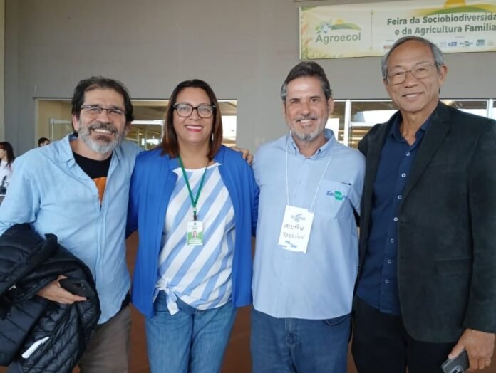 Four adults smile and pose together at an indoor event, wearing name badges, with a banner for Agroecol in the background.