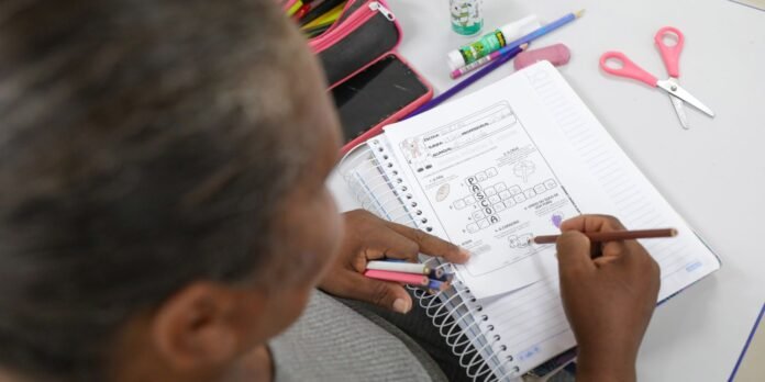 Student uses colored pencils to complete a worksheet in a spiral notebook on a desk with school supplies nearby, including a pencil case and scissors.