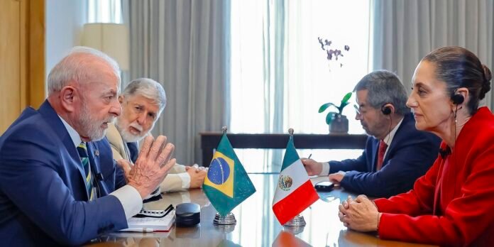 Four officials sit across a glass conference table, Brazil and Mexico flags between them during a formal meeting.