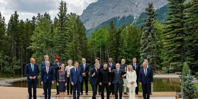 Group of world leaders posing for a photo on a wooden platform outdoors, with a dense forest and mountains in the background.