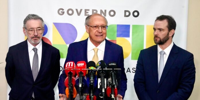 Three men in dark suits stand at a press conference, with many microphones in front of them.
