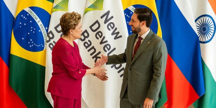 Two professionals shake hands in front of international flags and a banner reading 'New Develop Bank'.