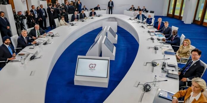 Global leaders sit around a white curved conference table on blue carpet, with a G7 France centerpiece display in the middle.