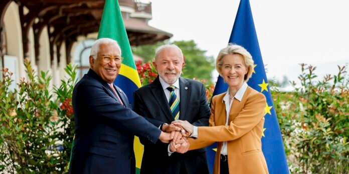 Three diplomats smile as they place their hands together in the center, with flags behind them.