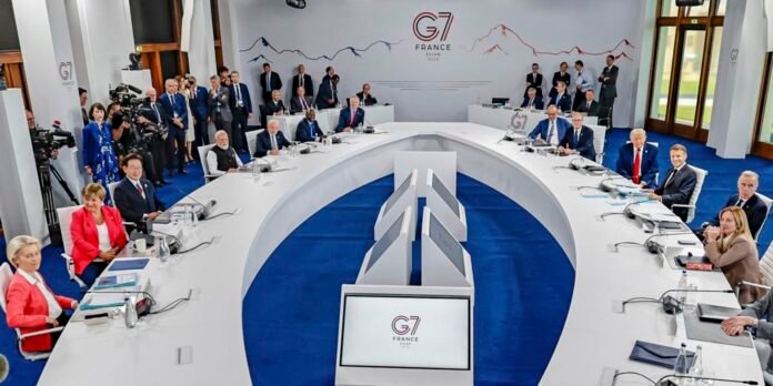 World leaders sit around a large white U-shaped conference table in a bright room with a G7 France banner and cameras watching from the sides.