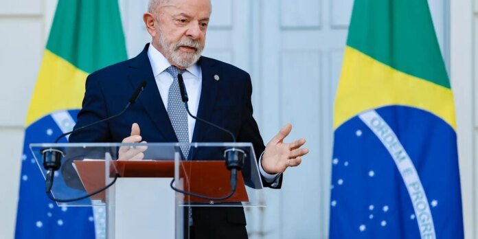 Older man in a dark suit and patterned tie speaks at a clear podium with two microphones, Brazilian flags in the background.