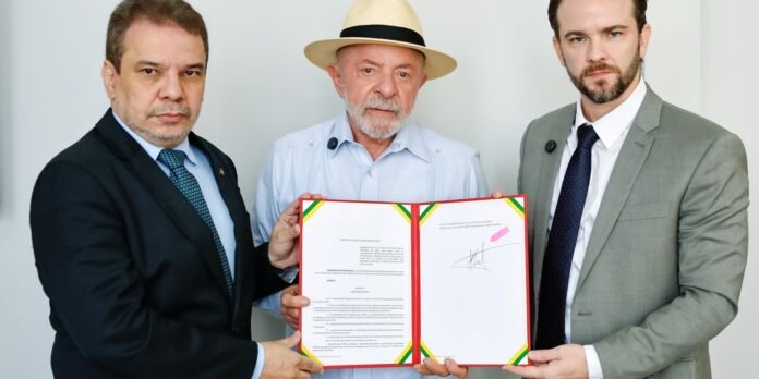 Three men in formal attire presenting and holding an open framed document with green-yellow-red corner accents, including a signature on the right page. The center man wears a straw hat and light shirt; two others in suits flank him, looking at the camera.
