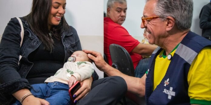 Smiling mother cradles a sleeping baby with a pacifier, baby in beige hoodie and jeans, in a busy room with a healthcare worker nearby.