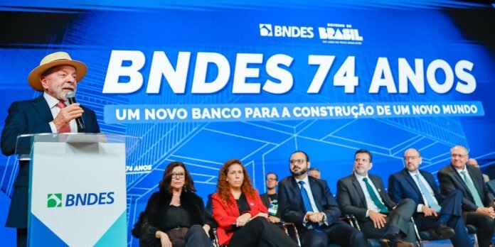 Speaker at a podium wearing a hat and suit, with a blue BNDES 74 Anos backdrop stating 'UM NOVO BANCO PARA A CONSTRUÇÃO DE UM NOVO MUNDO' and other panelists seated on stage behind him.