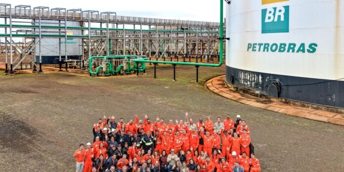 Group of Petrobras workers in orange coveralls posing for a photo in an industrial oil facility with green pipelines and a large white tank behind them.