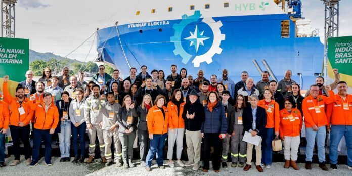 Group of diverse people in bright orange jackets posing for a photo in front of a large blue ship with a gear and star emblem at a port event.