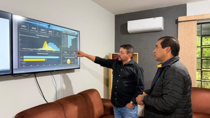 Two men in a meeting room review a large wall-mounted dashboard showing charts and metrics on a screen.