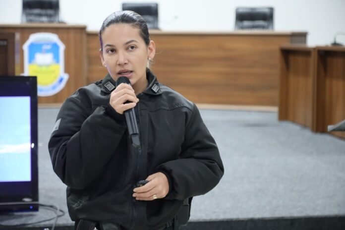 Woman in a black jacket speaks into a handheld microphone in a formal meeting room.
