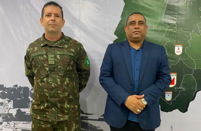 Two men stand side by side before a map backdrop: a soldier in camouflage with a Brazil patch beside a man in a blue suit and shirt.