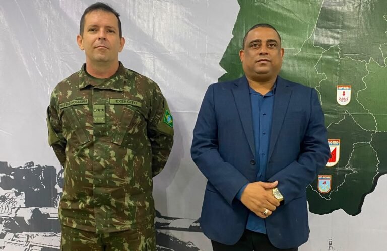 Two men stand side by side before a map backdrop: a soldier in camouflage with a Brazil patch beside a man in a blue suit and shirt.