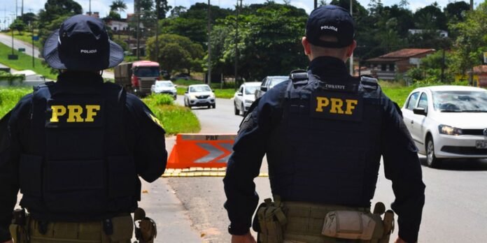Two PRF officers in tactical gear stand at a roadside checkpoint with an orange barrier and approaching cars on a sunny day.