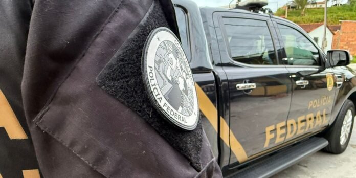 Close-up of a Polícia Federal badge on a dark uniform, with a black Federal Police pickup truck in the background.