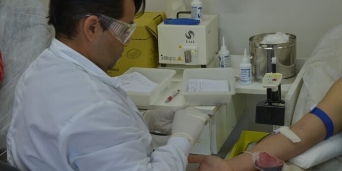 Phlebotomist in a white coat and gloves draws blood from a patient’s arm in a clinical setting with lab equipment nearby.