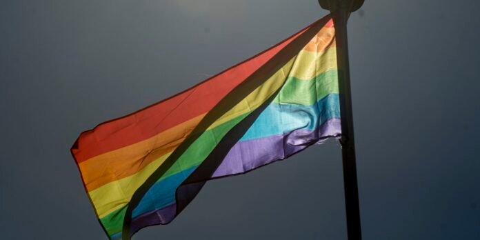 Rainbow Pride flag waving on a pole against a dark sky, representing LGBTQ+ pride with bright stripes from red to purple.