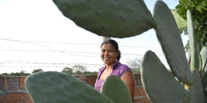 Smiling woman wearing a purple top stands among large prickly pear cactus pads outdoors, with a barbed-wire fence in the background.