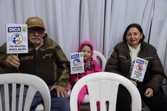 Three people sit side by side holding DICA signs—a man in a cap, a child in a pink hoodie, and a woman in a brown jacket.