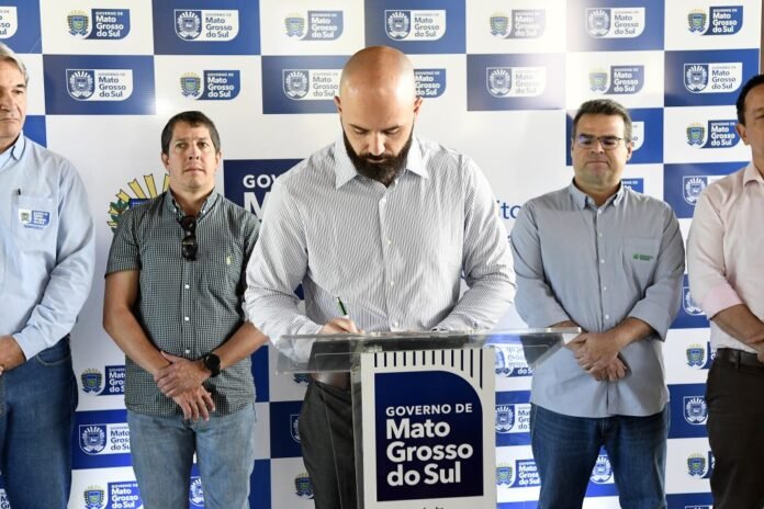 Man with a beard signs a document at a podium as others stand beside him during a press event with a backdrop displaying Mato Grosso do Sul government logos.