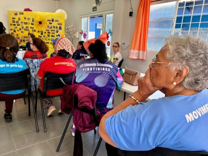 Elderly woman in a blue shirt sits at a table during a community workshop, with a bright yellow photo display in the background.