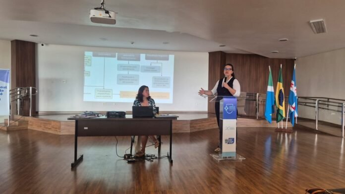 Female presenter at a blue podium giving a talk in a conference room, projection screen with a flowchart behind her, and a table with a laptop and projector in the foreground.