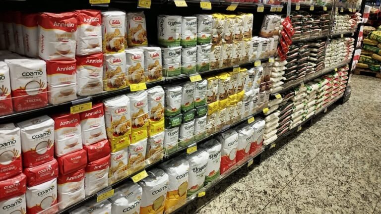 Aisle in a grocery store with tall stacks of large packaged bags (rice and flour) on shelves, in red, white, and yellow packaging with yellow price tags visible along the front edge of the shelves.